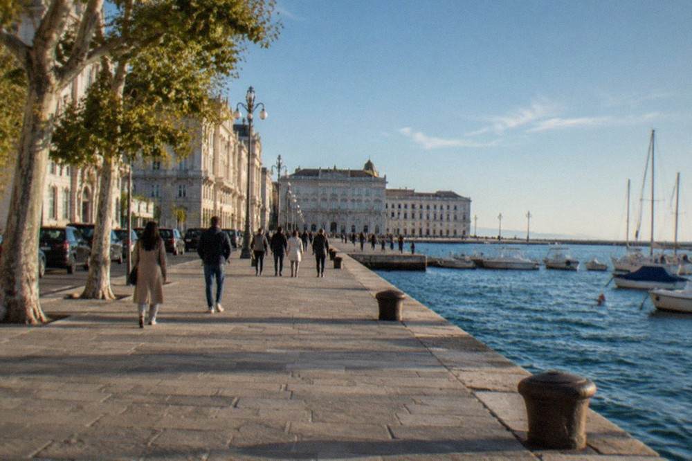 Passengers strolling along the Trieste seafront promenade toward a wide open square beside the sea