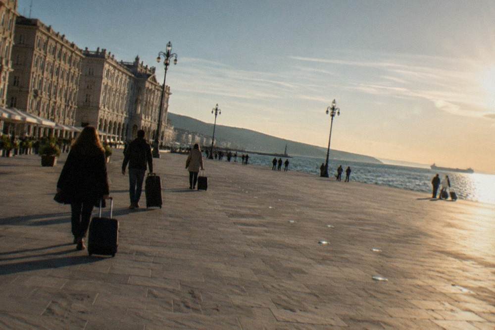 Passengers with suitcases walking along the seafront near Trieste's Piazza Unità d'Italia on a sunny morning