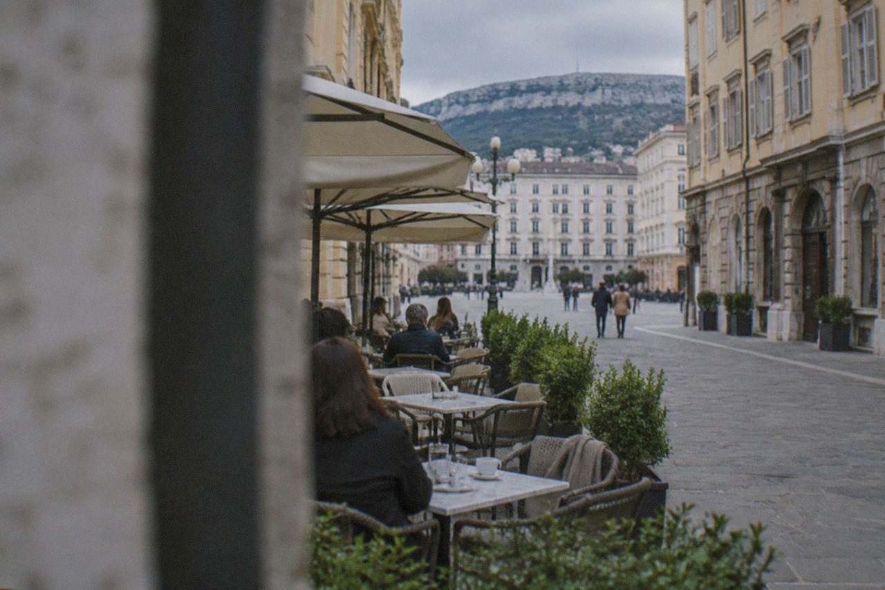 People at marble tables on a café terrace in Trieste beneath pale ochre stucco.