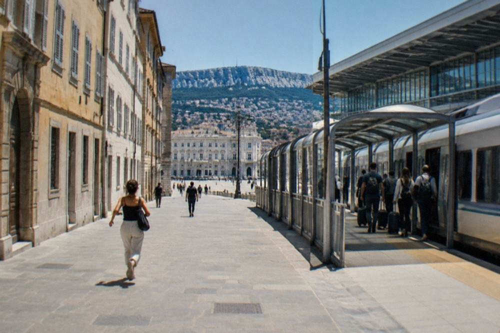 A regional train standing at a platform beside a modern airport terminal, with passengers boarding using a covered walkway.