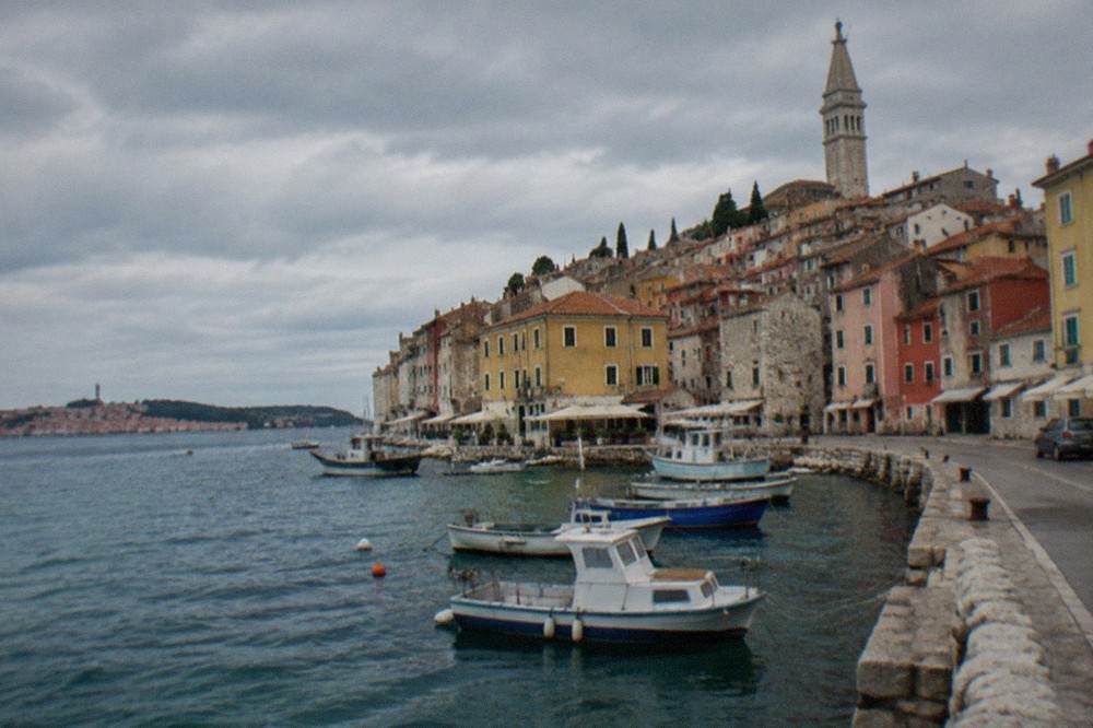 A coastal road above the Adriatic with red rooftops of a small border-town harbour in the distance