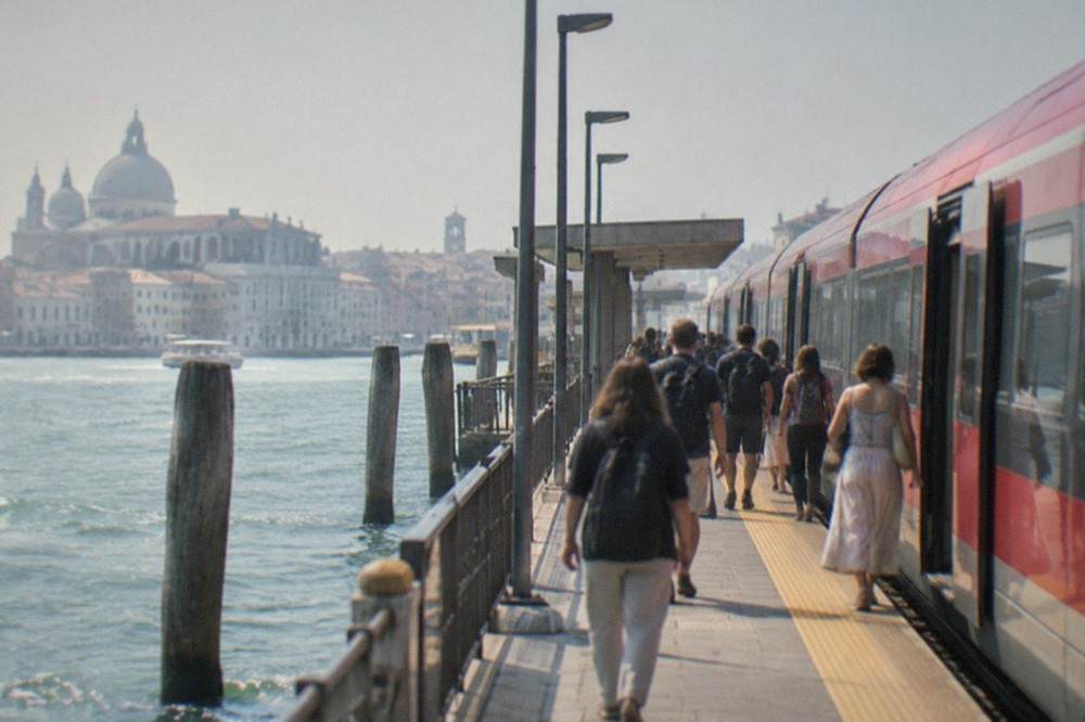 A red railway train waiting at a platform beside the seafront, with passengers boarding for a journey along the Adriatic coast