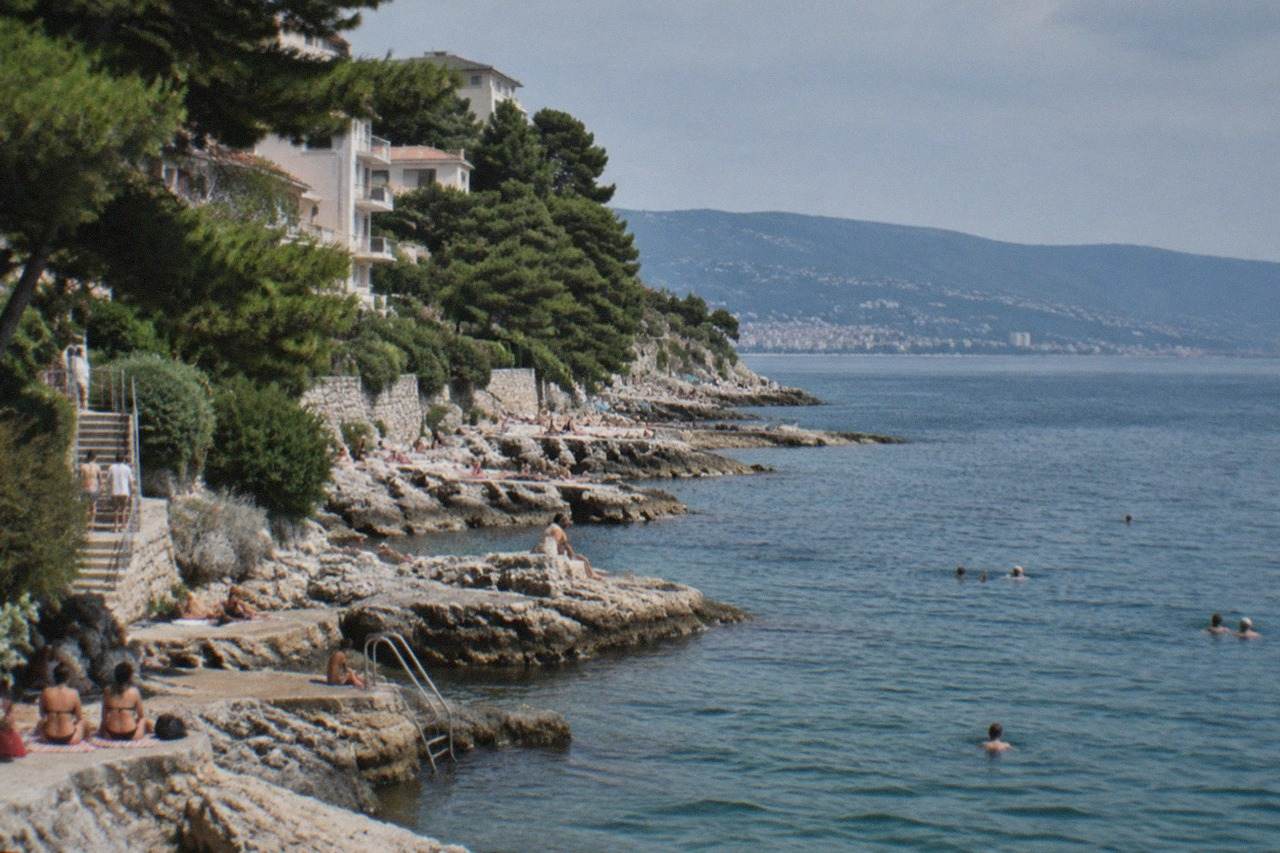 Swimmers and sunbathers along the rocky seafront at Barcola in Trieste, with the Adriatic gulf and distant hills behind