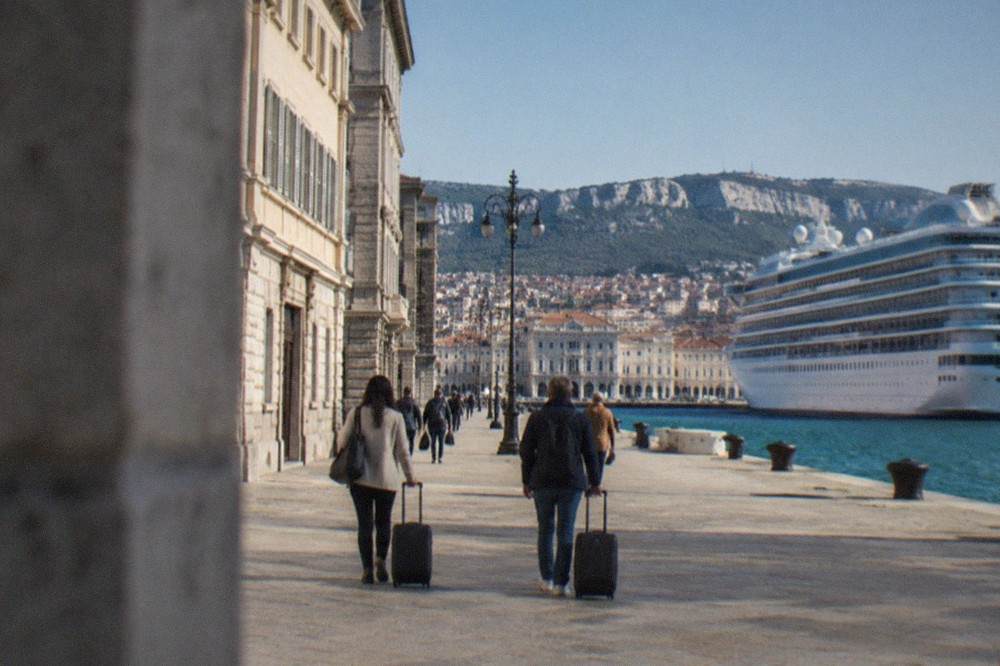 Passengers walking with luggage along the Trieste waterfront near the cruise terminal on a clear day