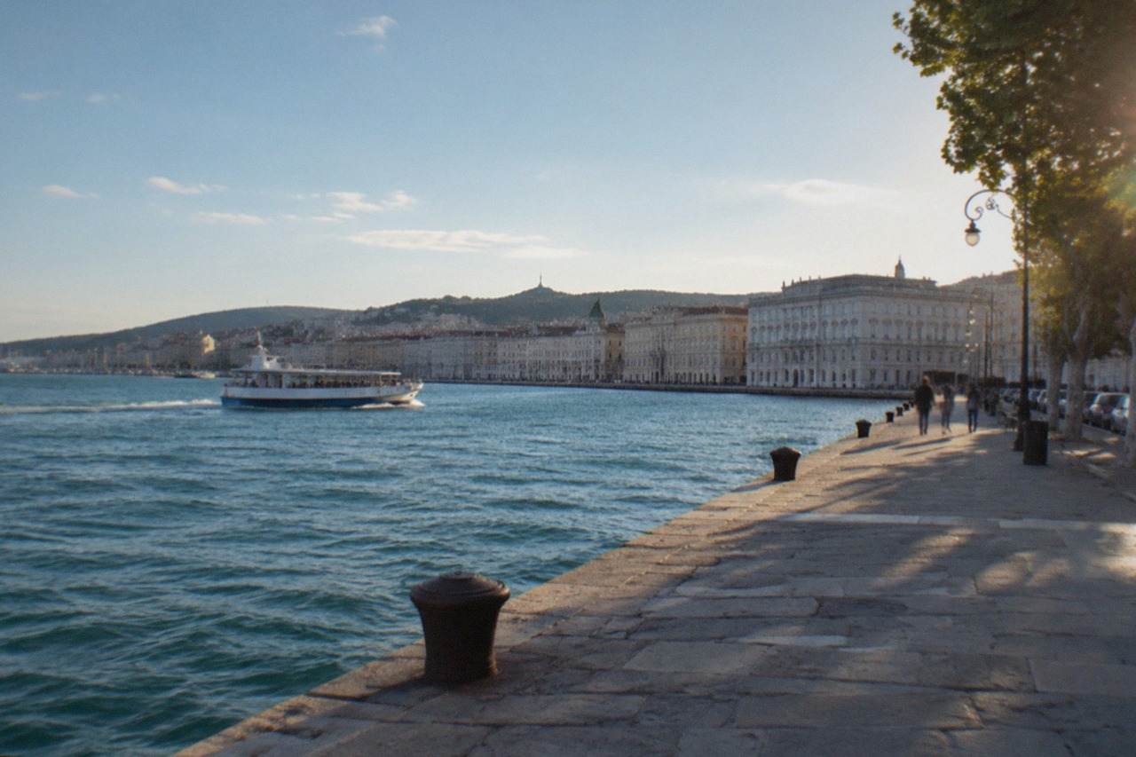 A small passenger ferry moving across the water of the Gulf of Trieste with the city's seafront facades behind it
