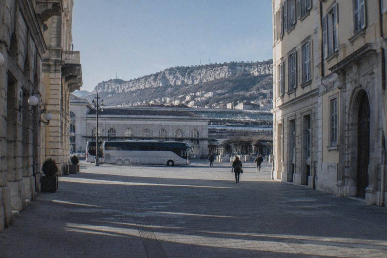 A long-distance coach parked at a city bus station with a railway station visible behind it