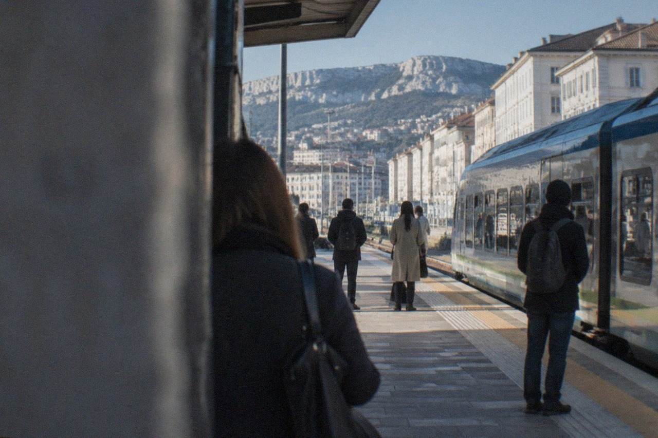 A train platform at Trieste Centrale station with passengers waiting beside a regional train on a bright morning