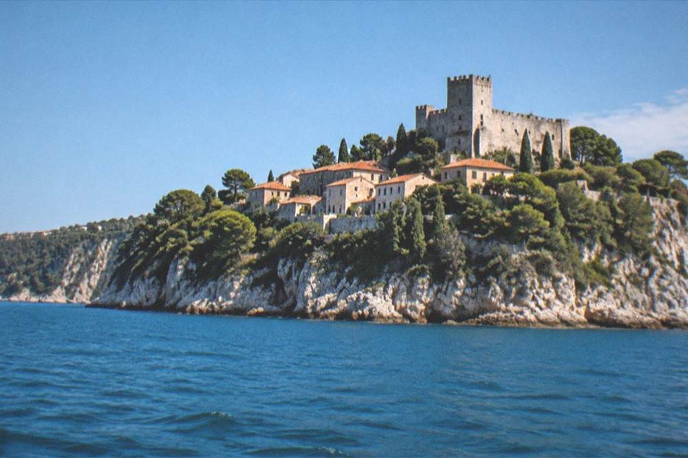 A wide view of a castle and village above a rocky Adriatic shoreline seen from the water