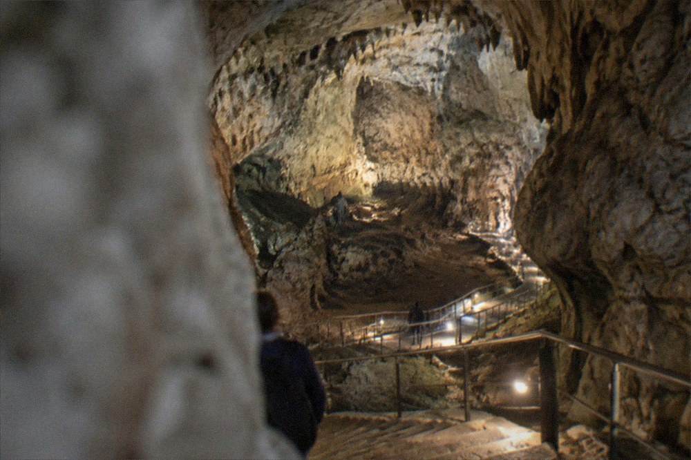 A vast limestone cave chamber lit by walkway lamps with a stairway descending inside