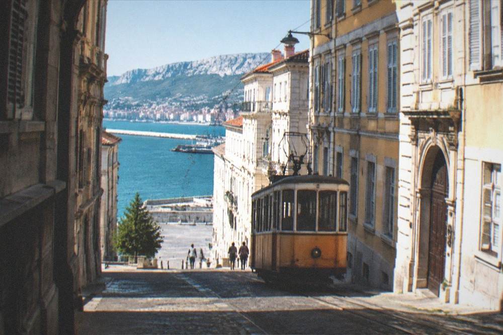 A vintage tram climbing a hillside street with the sea and a city visible far below