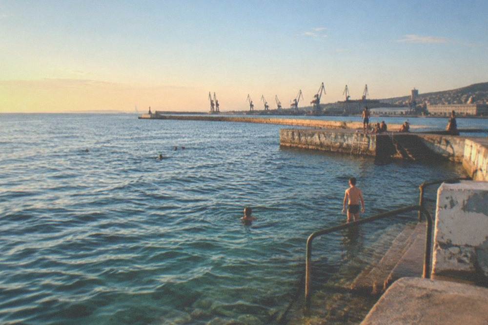 Children and adults swimming from a stone platform into clear Adriatic water on a sunny day