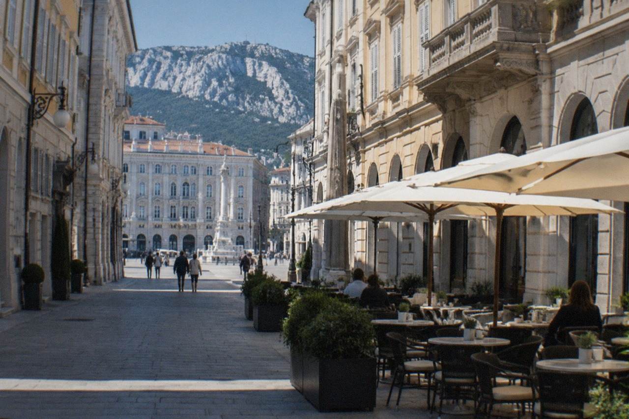 Passers-by cross a small bridge over the Canal Grande of Trieste, pale ochre facades along the pavements.