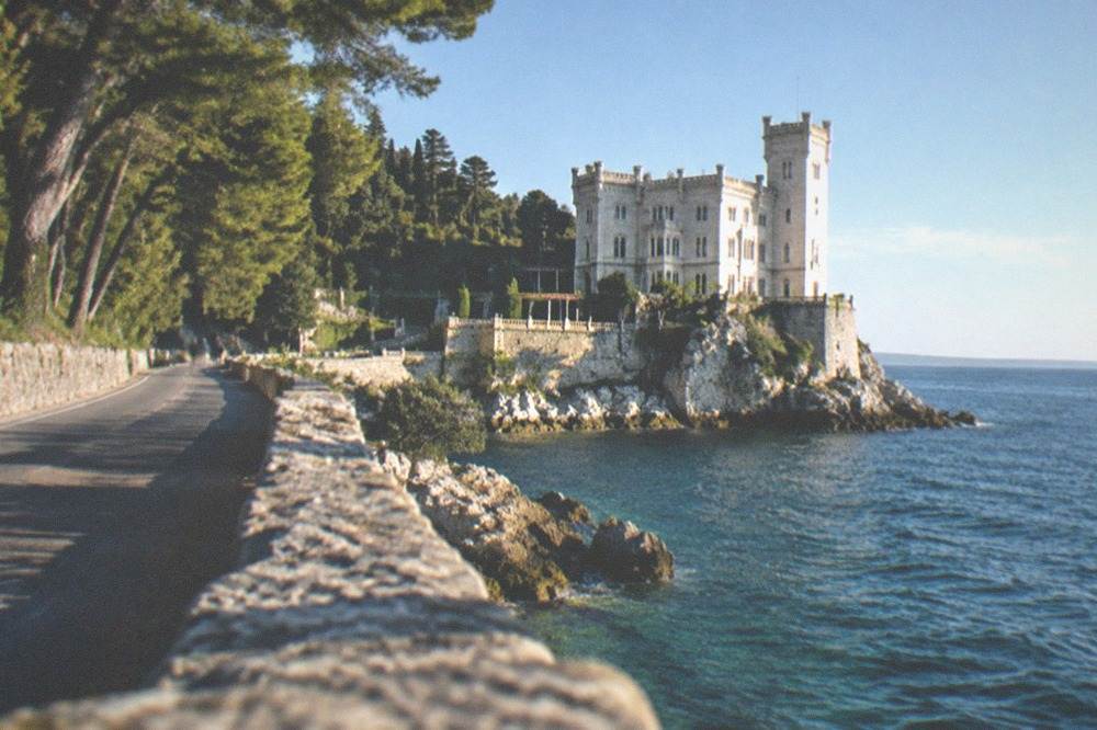 A coastal road with the sea on one side and trees on the other leading towards a castle headland