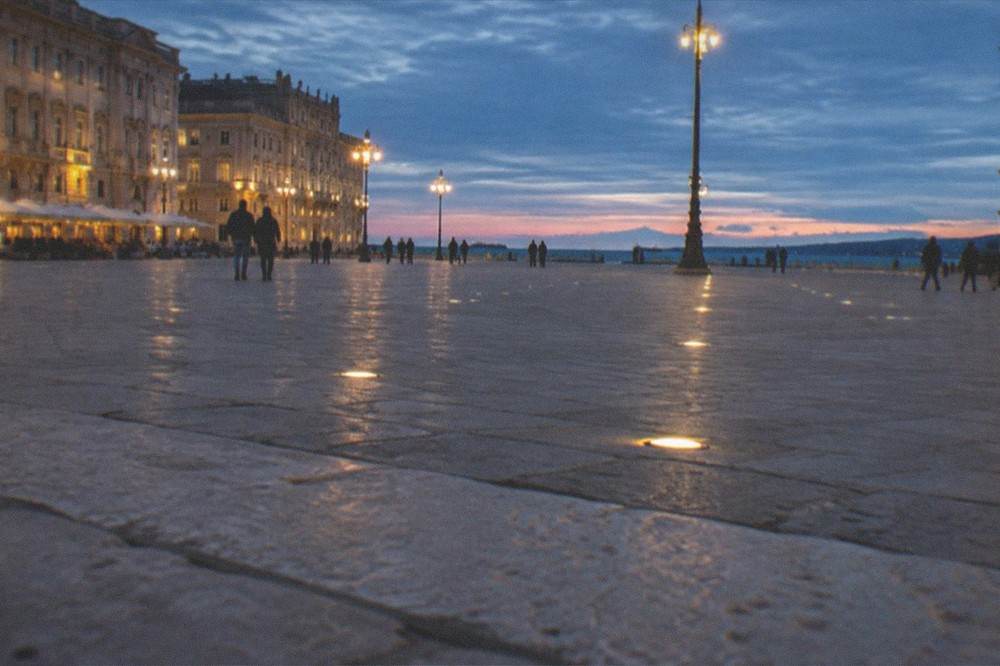 An Italian square at dusk lit by warm lamps with people walking near the waterfront