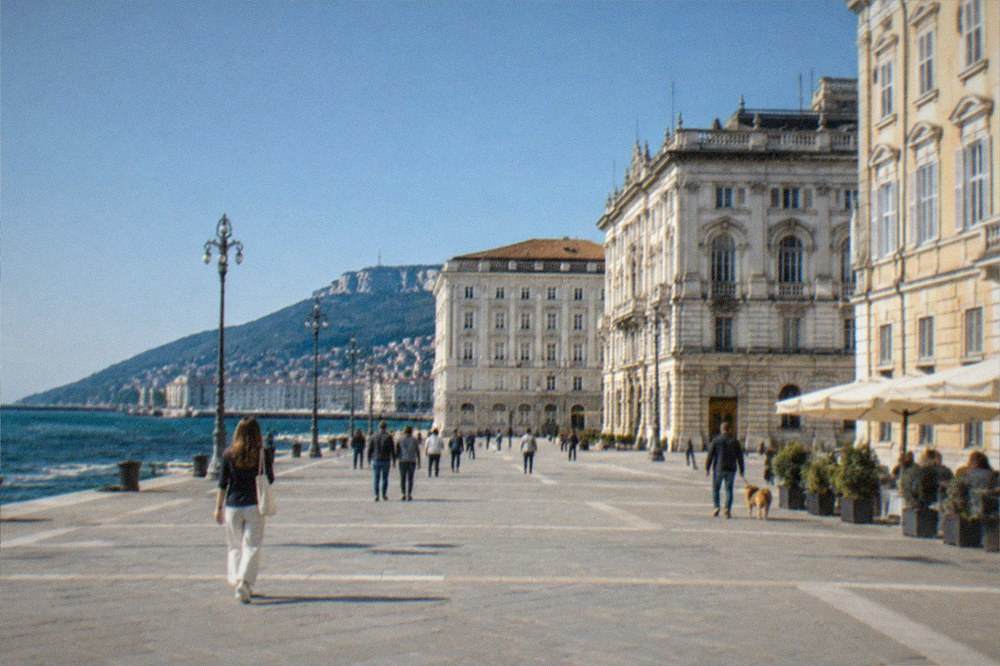 A wide sea-facing square in Trieste with ornate facades and people crossing the open paving