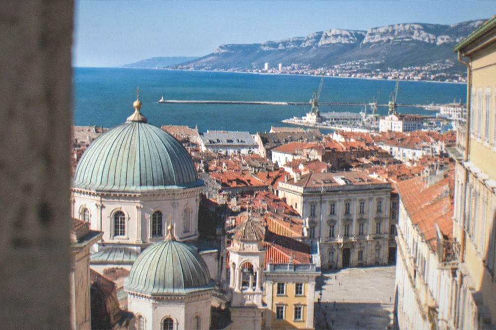 A wide view over Trieste's old town rooftops toward the sea with the Saint Spyridon church domes in the foreground