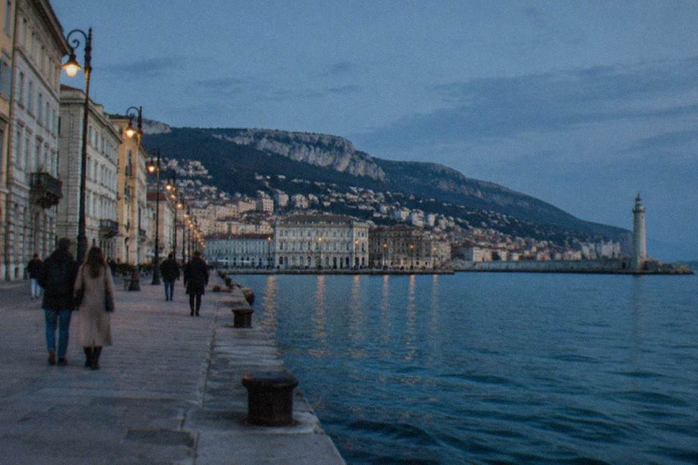 Walkers on the Trieste Rive promenade with the lighthouse visible along the shoreline