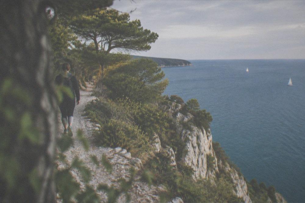 A cliff path in warm evening light above a calm blue sea with sailboats in the distance
