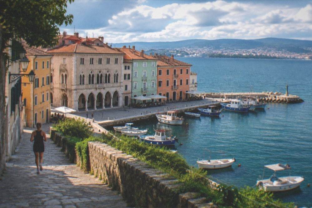 A hillside path above a blue bay with a town and distant city across the water