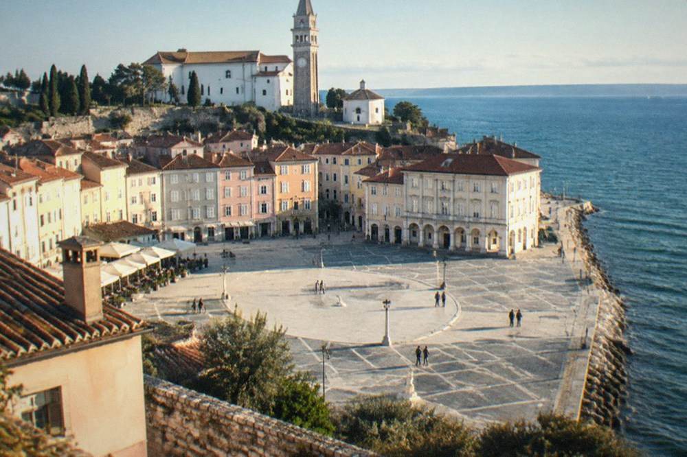 A broad stone-paved square in a coastal town with pastel-coloured buildings around the edge.