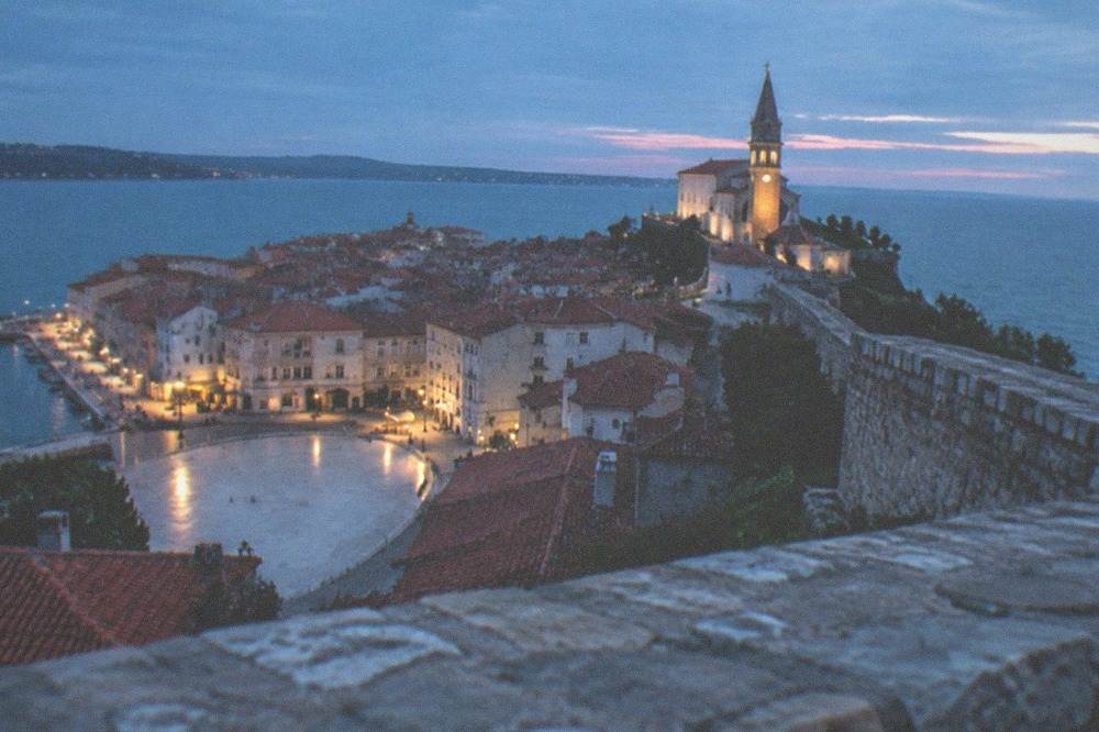 A stone defensive wall above a coastal old town, with the sea and distant coastline beyond.