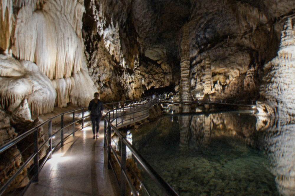 A lit underground walkway winding past pale rock formations reflected in a still pool