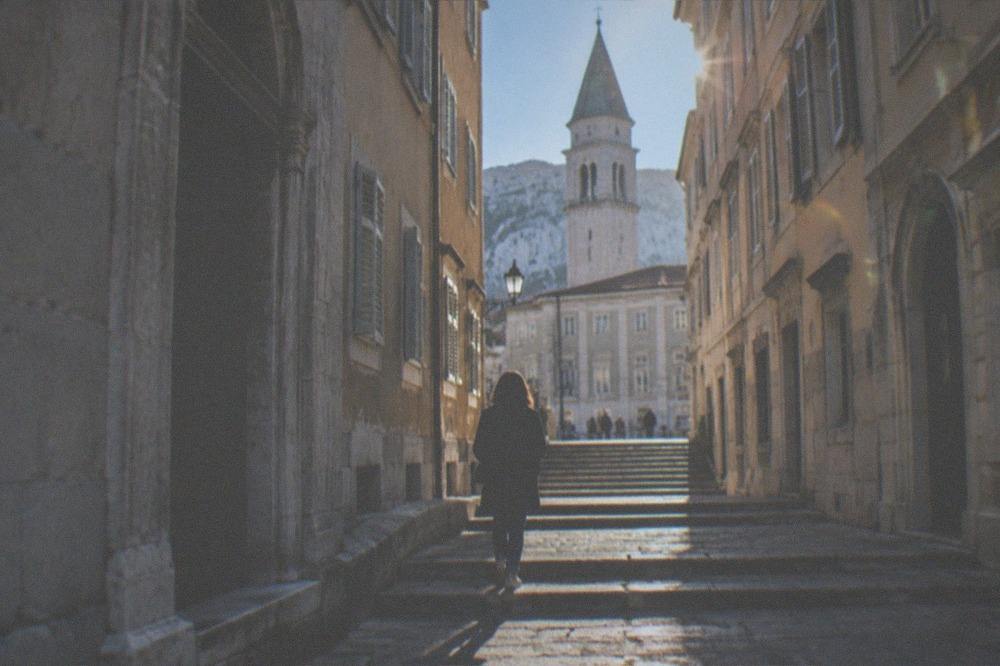 A visitor walking alone up a stepped lane in the old town of Trieste towards a cathedral bell tower