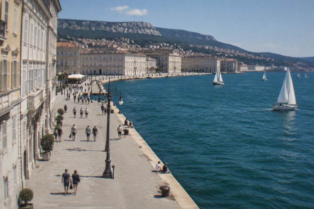 People strolling along the Rive seafront in Trieste on a bright afternoon with sailboats on the water