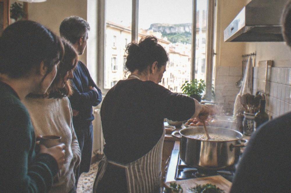A home kitchen in Trieste with a cook stirring a large pot of soup while a small group watches