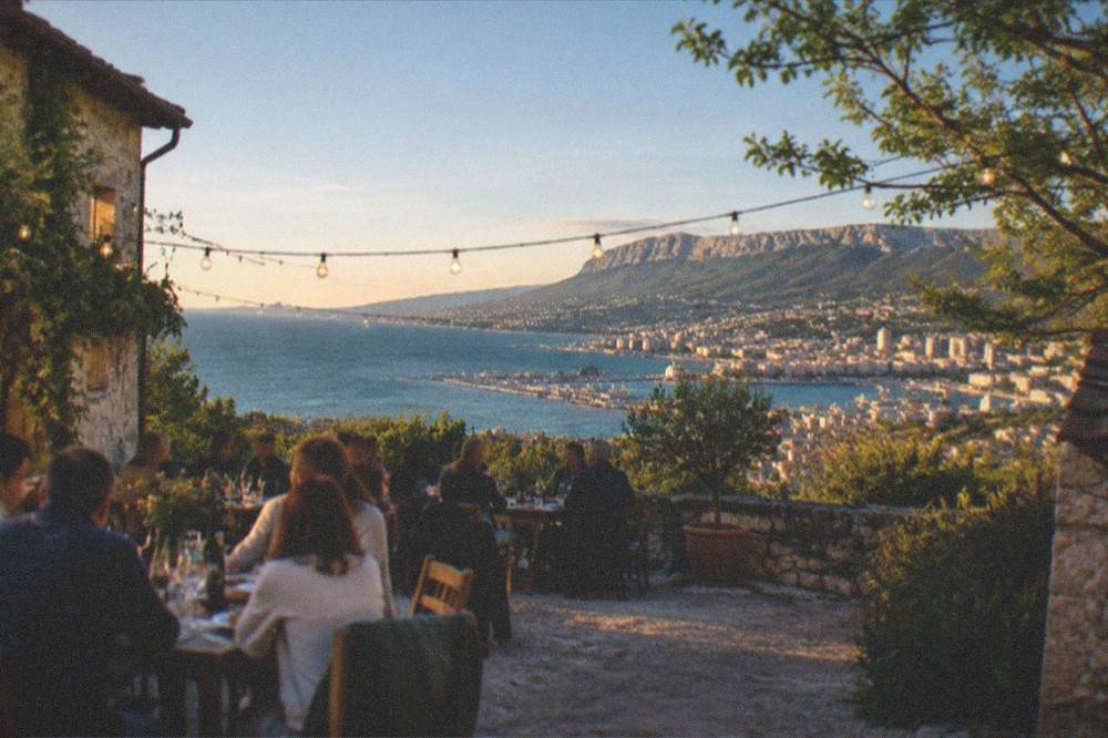 A farmhouse courtyard at dusk with string lights, full tables and the lights of Trieste below on the coast