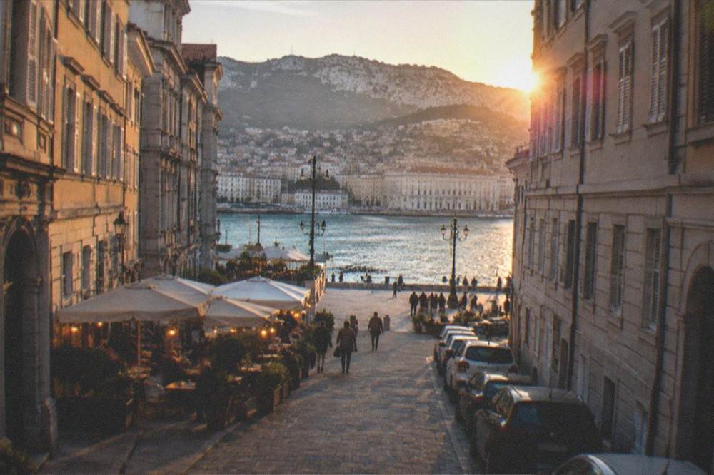 An evening street in an Italian port city with lit cafe terraces and parked cars along the kerb