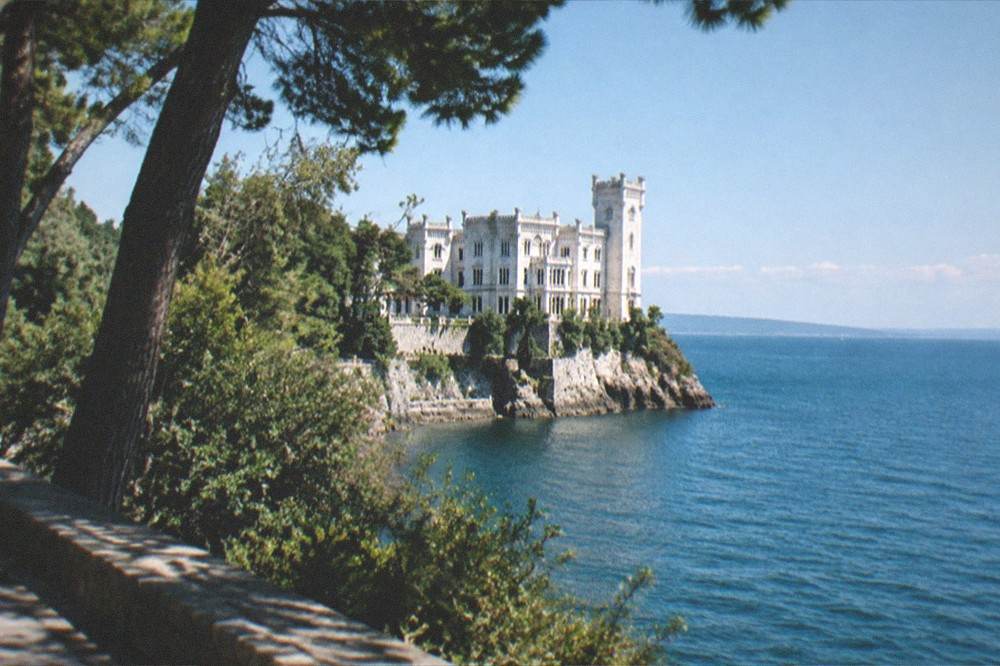 A white castle on a promontory above the sea seen from a coastal road with trees in the foreground