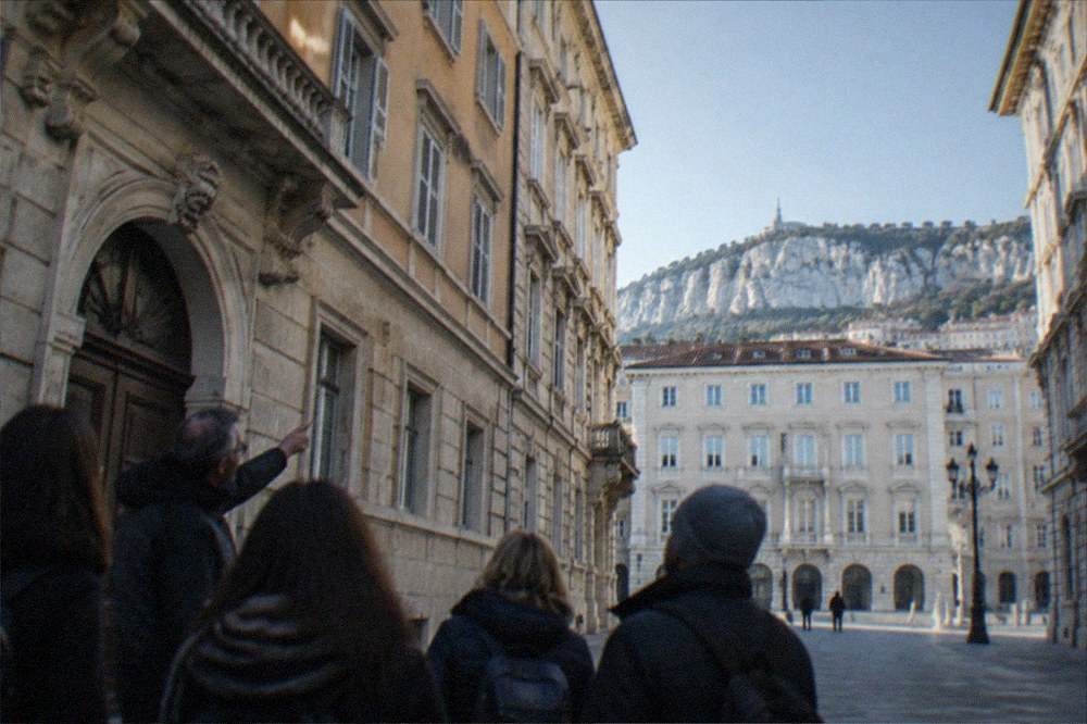 A local guide pointing out details on a building facade to a small group in a Trieste square