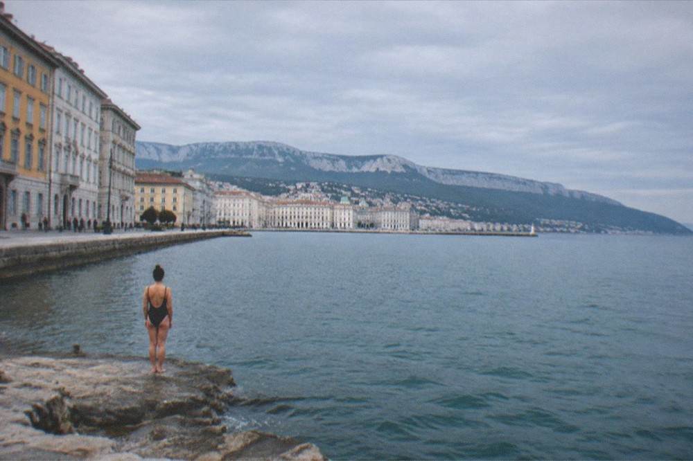 A swimmer standing on a rock platform looking out over the calm blue water of the Gulf of Trieste