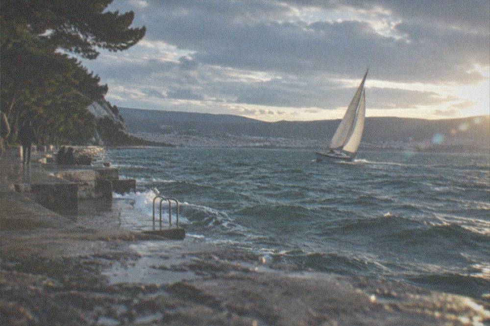 A yacht heeling under full sail in open water with a hilly coastline and a distant city skyline behind it