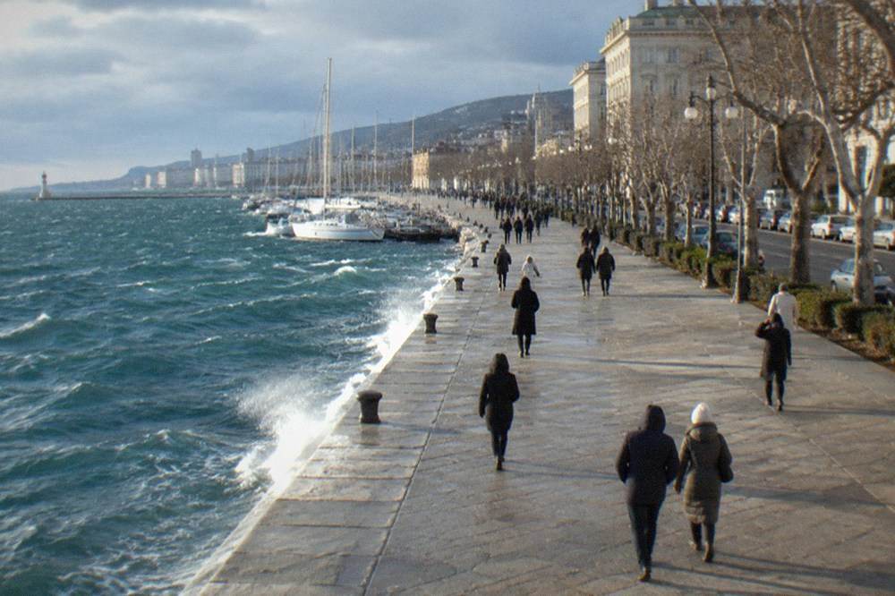 People in winter coats walking along a seafront promenade under grey skies in Trieste