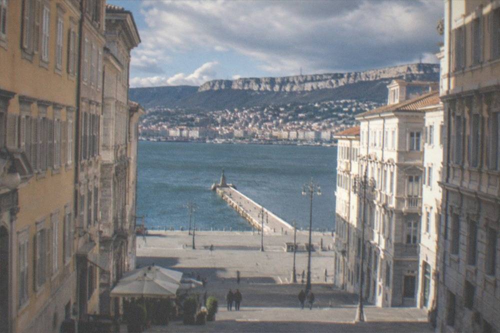 A stone pier reaching into a calm blue gulf with a city skyline behind it