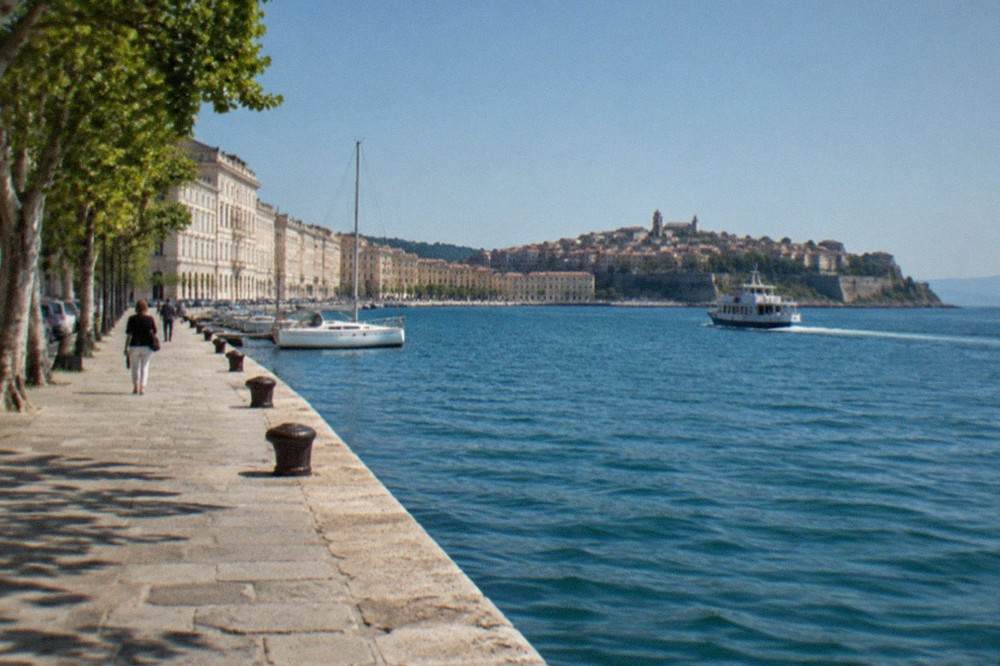 A small passenger ferry crossing calm blue water toward a walled coastal town