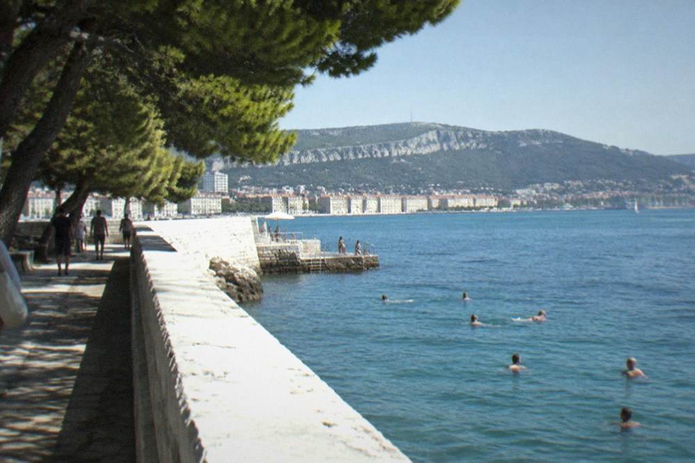 A high white wall running out into the sea at a bathing establishment, with swimmers in the water on one side